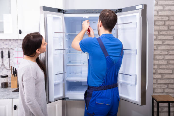 Technician in blue uniform repairing a double-door refrigerator while a woman observes in a modern kitchen.