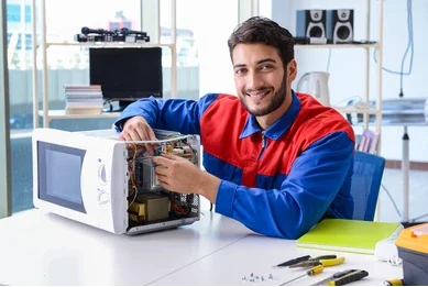 Smiling technician in red and blue uniform repairing a microwave oven on a desk with tools.